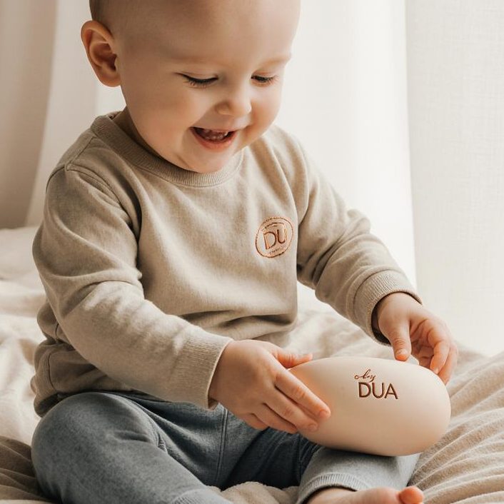 Babyprodukte von by DUA Hand hält einen runden, rosa Silikonlöffel und ein Spielzeug mit Hasenohren.