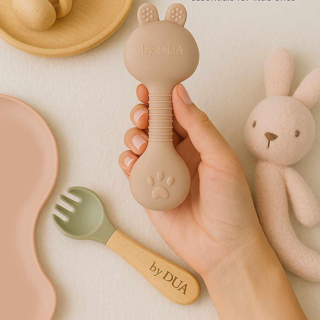 Babyprodukte von by DUA Hand hält einen runden, rosa Silikonlöffel und ein Spielzeug mit Hasenohren.